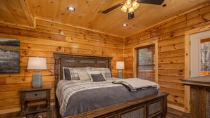 Un dormitorio de cabaña de madera con una cama, dos mesitas de noche, un ventilador de techo y una ventana con persianas.