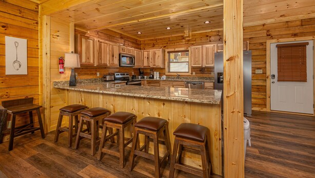 Interior de una cabaña de madera con una zona de cocina que incluye encimeras de granito, taburetes de madera y una barra.