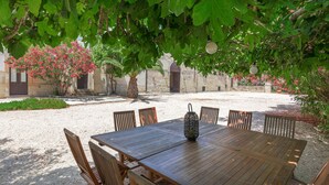 Una mesa de madera al aire libre con sillas, rodeada por un área de grava con árboles y un edificio al fondo.