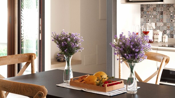 Un comedor con una mesa negra, sillas de madera y dos jarrones con flores moradas. Hay una cesta de frutas sobre la mesa.