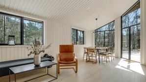 Una habitación amplia con piso de madera, un sillón de cuero, una mesa de comedor con sillas y ventanas grandes que ofrecen vista a árboles.