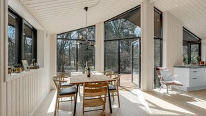 Un comedor amplio con una gran mesa y sillas, ventanas grandes que ofrecen vistas de los árboles circundantes y una cocina con un fregadero y mostrador.
