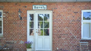 Ein Backsteinhaus mit einer weißen Tür und einem Schild, auf dem "Havely" steht. Vor den Stufen stehen zwei Blumentöpfe und links daneben eine Bank.