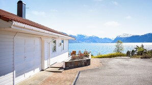 Una casa con puerta de garaje, un pequeño porche con banca y vista a un lago con montañas al fondo.