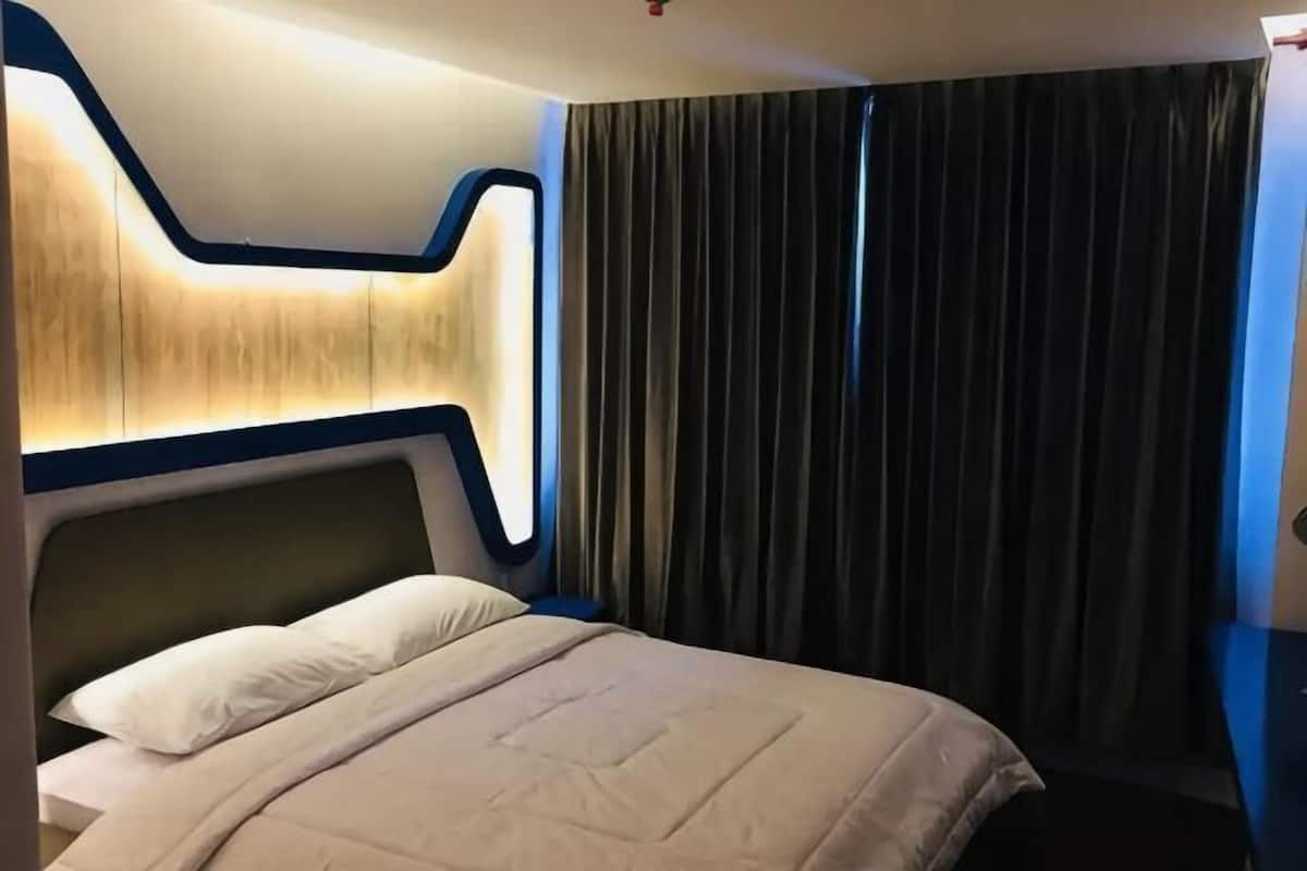 Una habitación de hotel con una cama, un cabecero con diseño iluminado, una cortina oscura y una lámpara de pared.