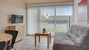 Ett modernt vardagsrum med en platt-TV, en grå soffa, ett matbord i trä med stolar och en skjutdörr i glas som leder ut till ett utomhusområde.