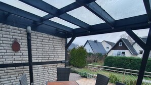 An outdoor patio with a wooden table and wicker chairs, a brick wall, and a view of houses in the background. - Vacation home Weserbergland, for up to 8 persons (Höxter)