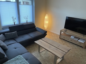 Un salon moderne avec un canapé d’angle, une table basse en bois, un téléviseur posé sur un meuble en bois et un grand vase.