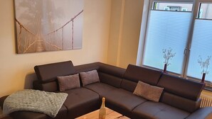 A modern living room with a dark brown sectional sofa, a wooden coffee table with a bottle and candle, and a large abstract painting on the wall. - Vacation home Weserbergland, for up to 8 persons (Höxter)