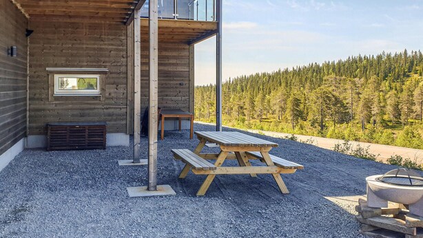 A wooden cabin with a balcony overlooking a forest. There is a picnic table and a fire pit outside.