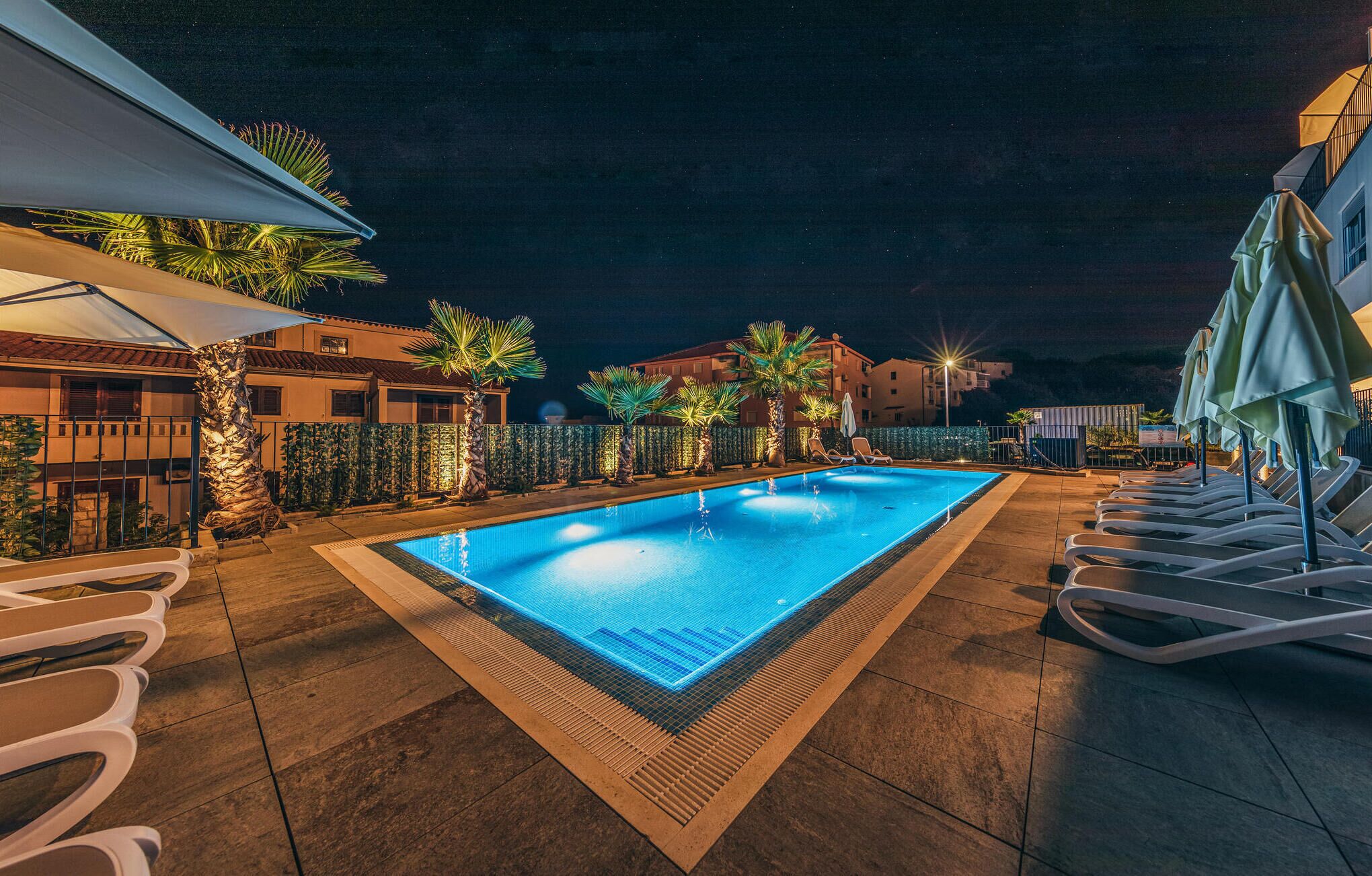 Ein Pool bei Nacht mit glatter, blauer Wasseroberfläche, umgeben von Liegestühlen und Palmen, mit Gebäuden im Hintergrund.