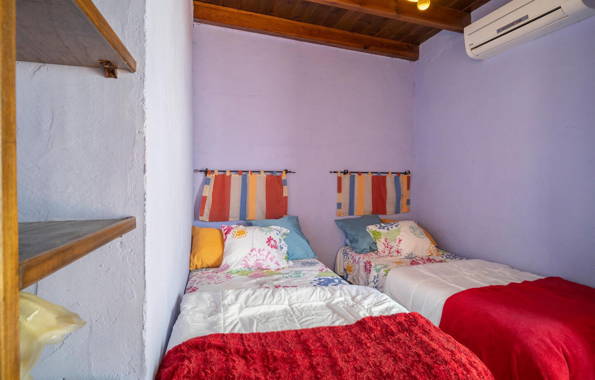 A small, cozy room with two beds, each with colorful bedding and striped curtains. There is a wooden shelf and a red blanket on one bed.