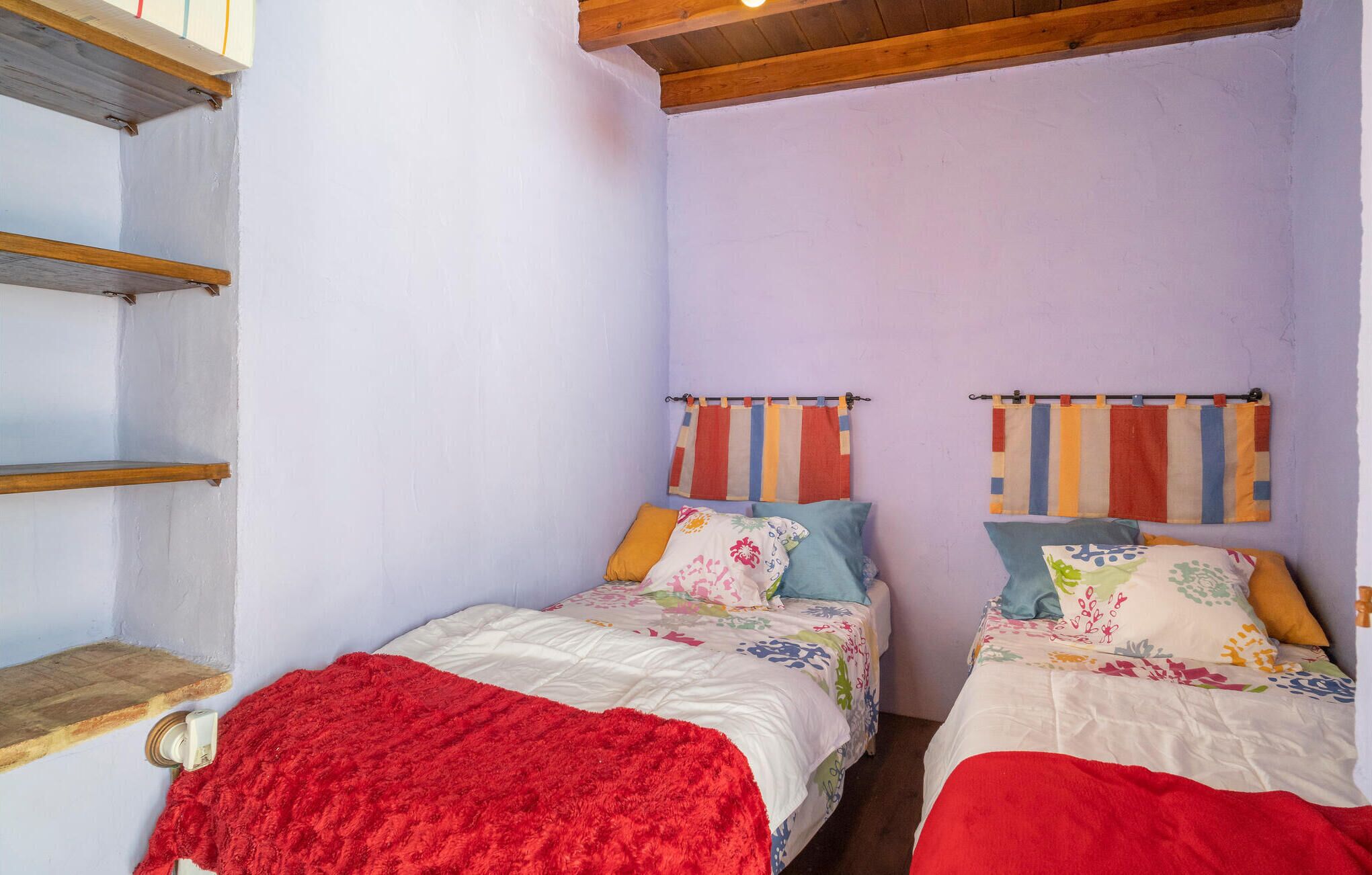 Une petite chambre à coucher accueillante, avec deux lits simples, une couverture rouge sur le lit de gauche et des rideaux à rayures aux fenêtres.
