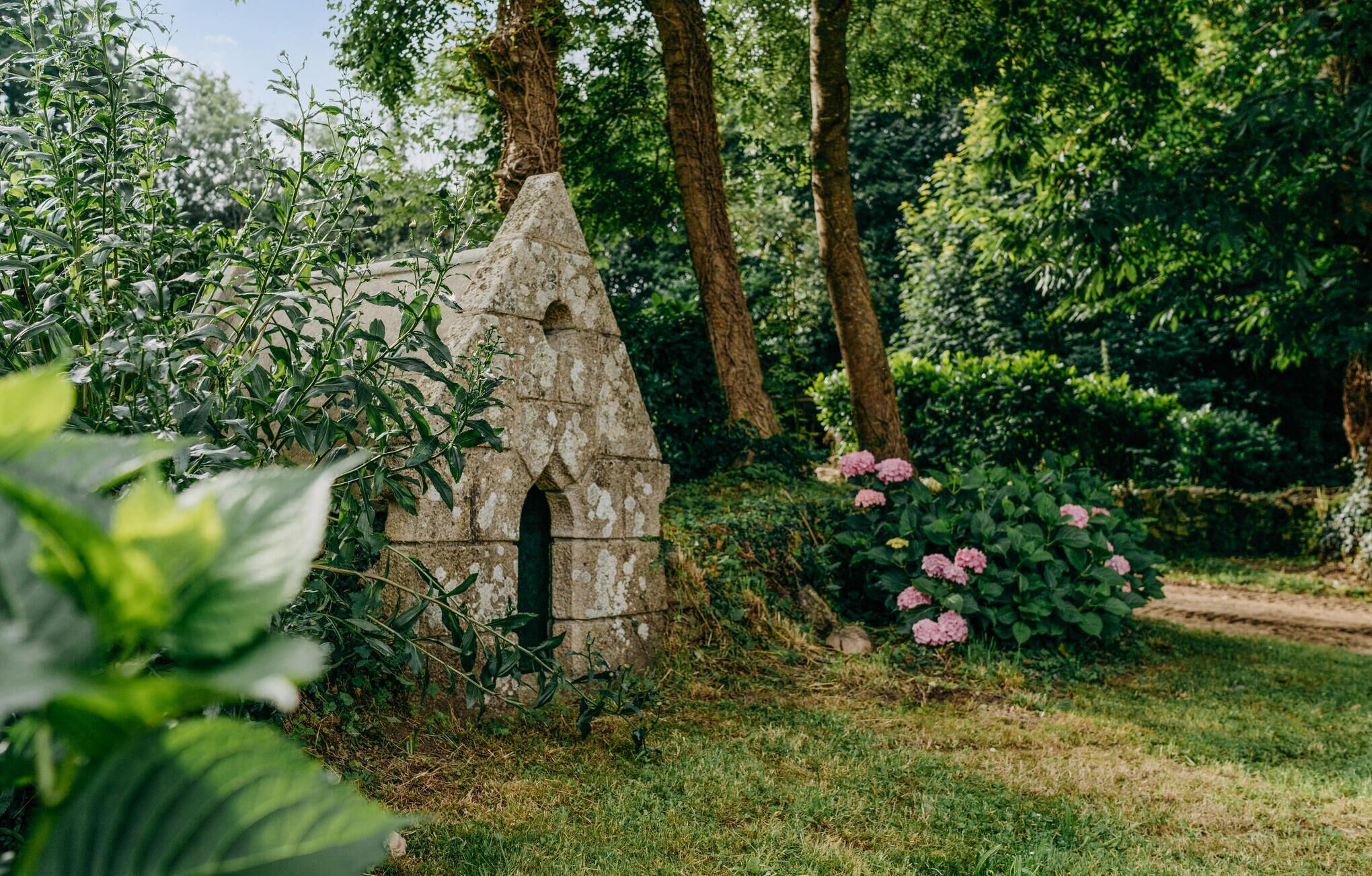 A stone structure with an arched doorway, surrounded by lush greenery and pink flowers.