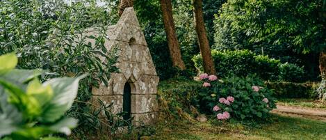 A stone structure with an arched doorway, surrounded by lush greenery and pink flowers.