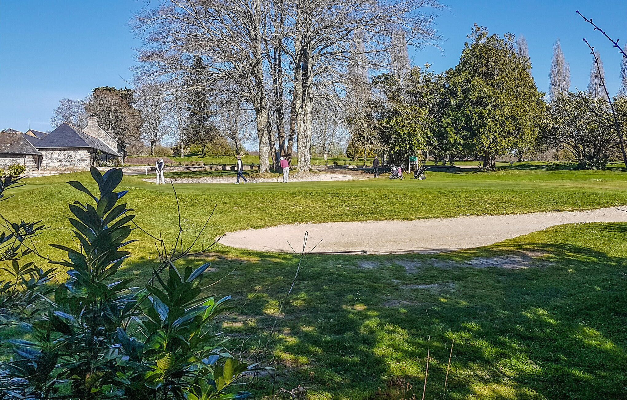 Un parcours de golf avec des bunkers de sable, des arbres et un bâtiment en pierre en arrière-plan.