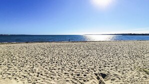 A sandy beach with footprints leading to the water, a clear sky, and a distant shoreline.