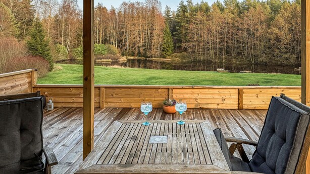 Een houten terras met tafel en stoelen, uitkijkend op een grasveld en bos.