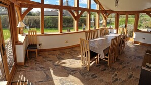 A dining area with a large table and chairs, wooden beams, stone flooring, and large windows offering a view of a garden and neighboring houses.