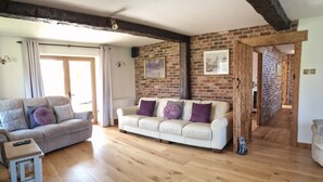 A living room with a white sofa, a smaller sofa, wooden flooring, a brick wall, and a ceiling with exposed beams.