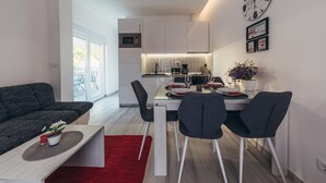 A modern kitchen with a microwave, stove, and sink. There is a dining area with a table set for two, two chairs, and a red rug. A clock and framed pictures are on the wall. - Nice apartment in Okrug Gornji (Okrug Gornji)