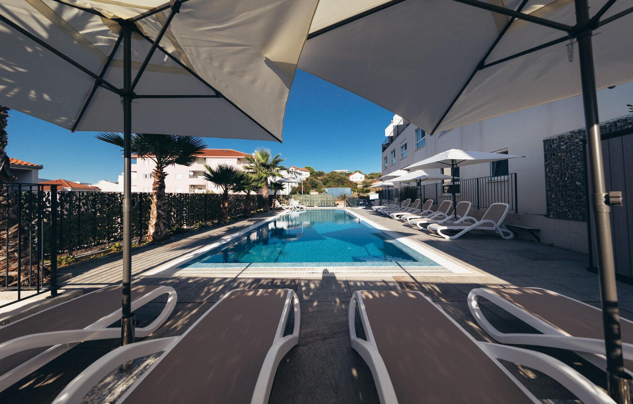 A swimming pool with lounge chairs and umbrellas, surrounded by a fence and palm trees.