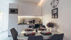 A modern kitchen with white cabinets, a built-in oven, and a dining area with a glass table set for four. - Nice apartment in Okrug Gornji (Okrug Gornji)