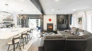 Um espaço de estar moderno com lareira central, área de cozinha e zona de estar com sofá e banquetas.