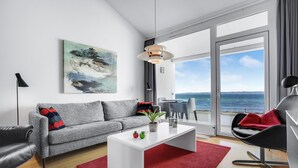 Een moderne woonkamer met een grijze bank, een witte salontafel en een rood vloerkleed. Aan de muur hangt een schilderij en door grote ramen is er uitzicht op zee.