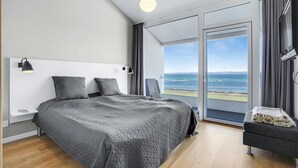 Een moderne slaapkamer met een groot bed, twee bedlampen, een nachtkastje, een stoel en uitzicht op de oceaan door een schuifdeur.