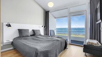 Et moderne soveværelse med en stor seng, to sengeborde, lamper, en stol og havudsigt gennem en skydedør.