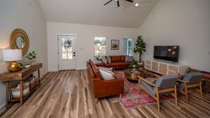 Un salón espacioso con un sofá de cuero marrón, una mesa de centro de madera y un televisor sobre un mueble blanco.