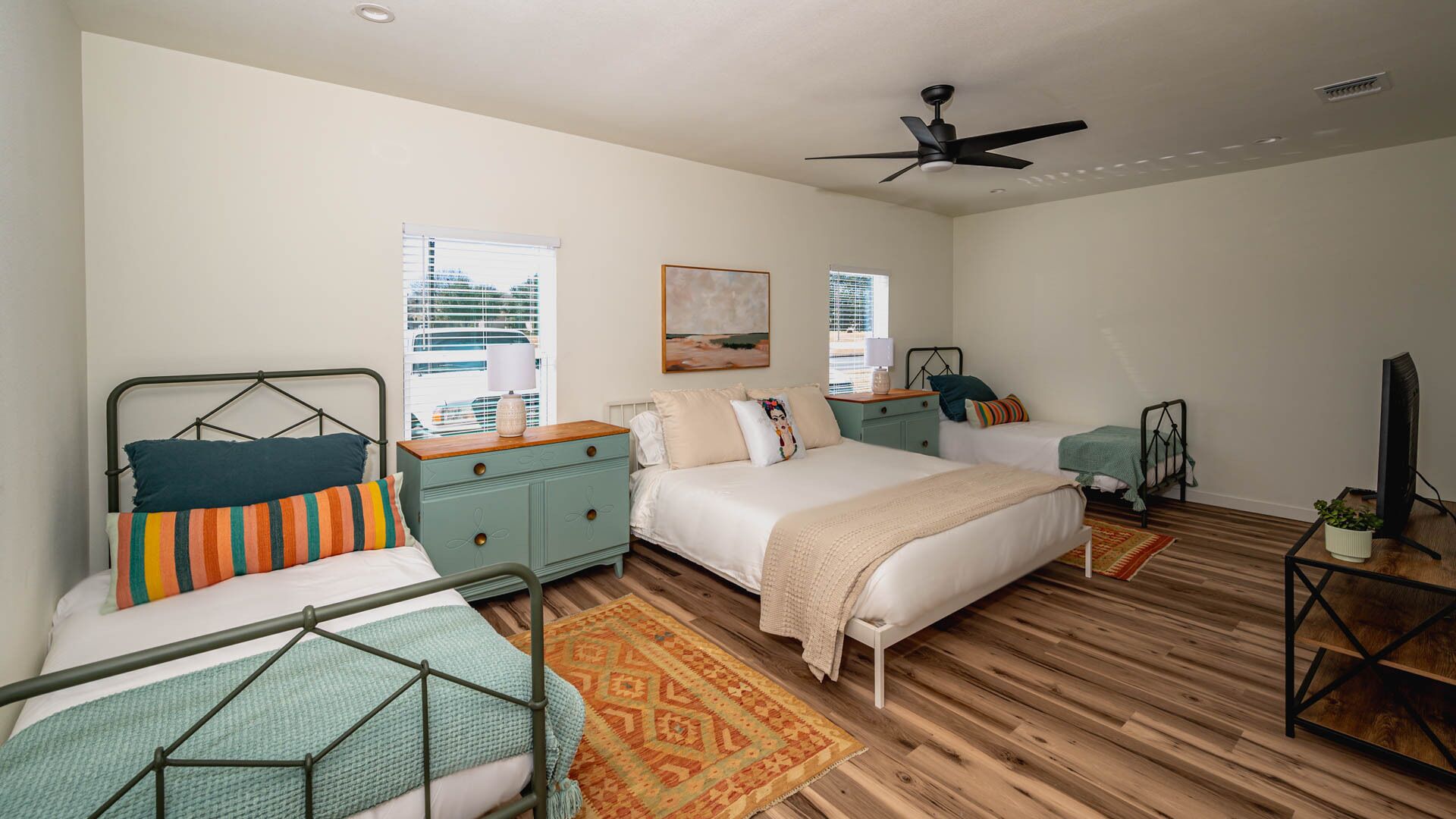 A bedroom with a bed, two bedside tables, a TV, a ceiling fan, and a window with blinds.