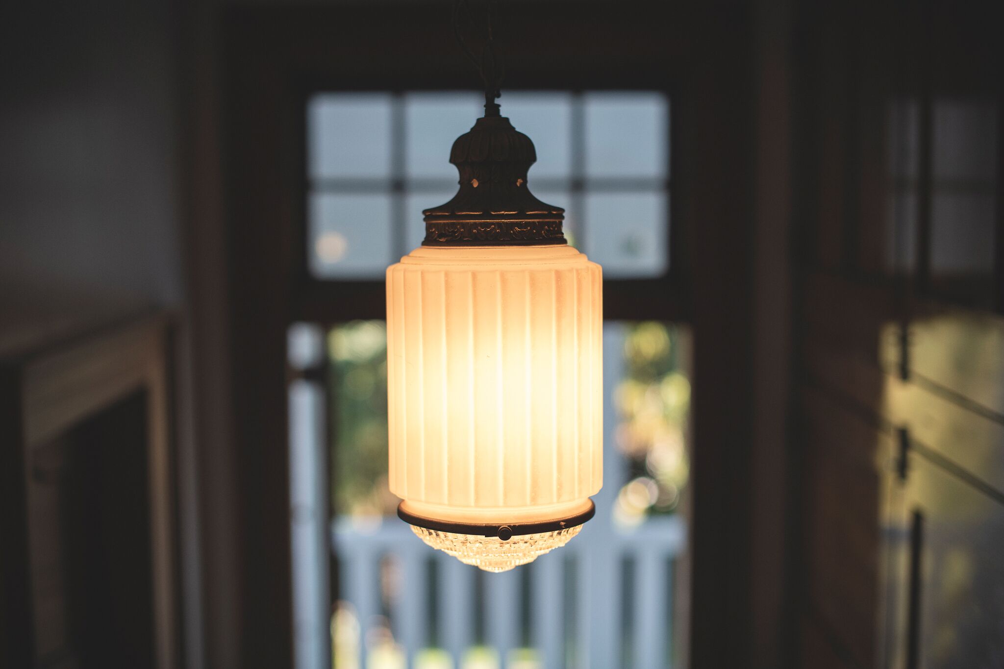 A hanging light fixture with a ribbed design and a decorative top, suspended in a room with a window in the background.