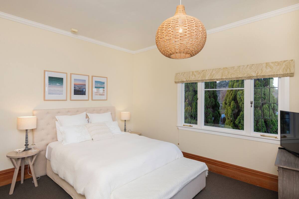 Una cama bien tendida con sábanas blancas, una lámpara redonda de fibras naturales encima y una ventana con vistas a árboles.