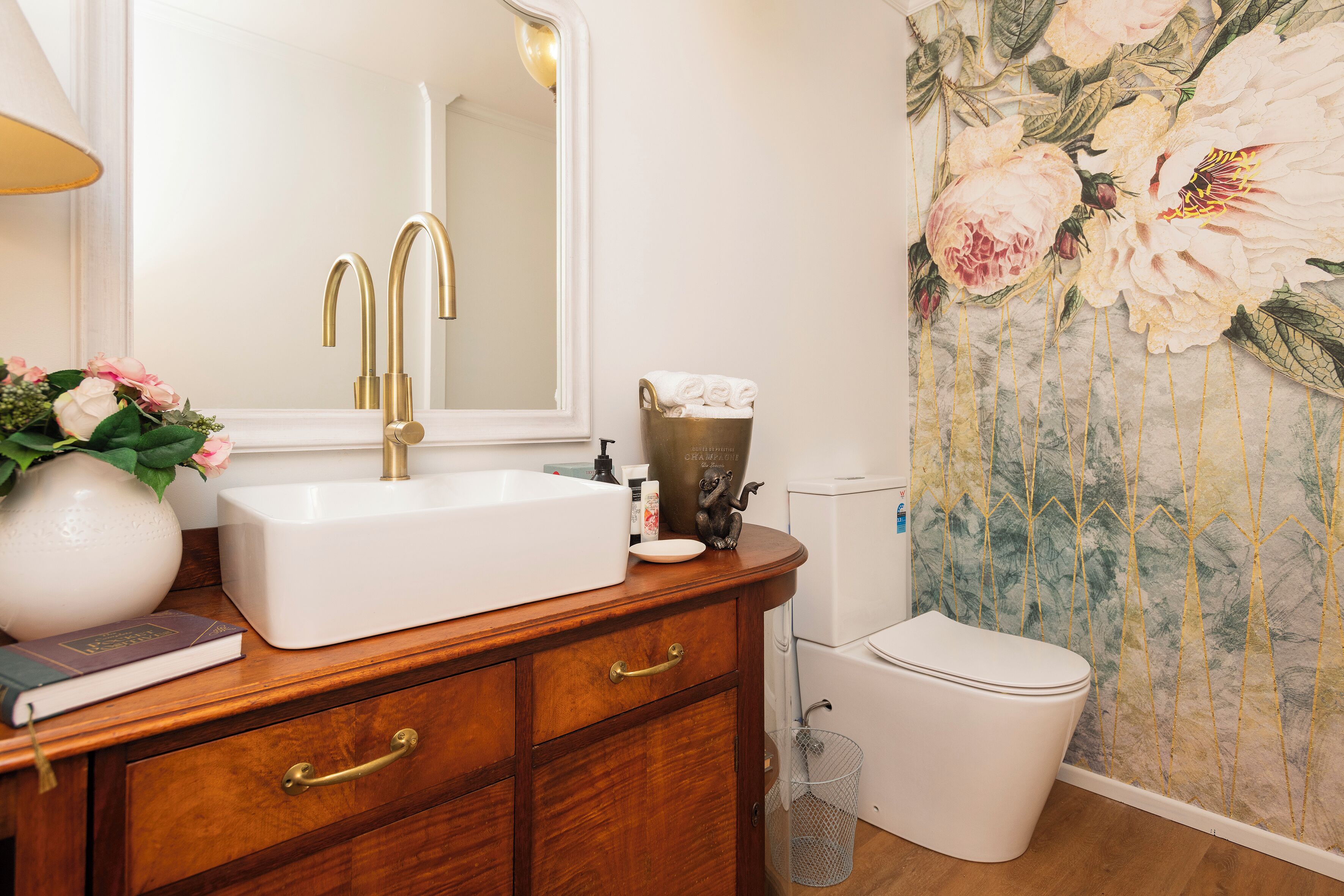 Un baño con lavamanos blanco, grifería dorada, un espejo, inodoro y un revestimiento floral decorativo.
