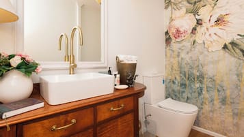Un baño con lavamanos blanco, grifería dorada, un espejo, inodoro y un revestimiento floral decorativo.