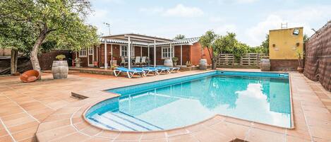 Una piscina rectangular con solárium empedrado, rodeada de sillones de playa y una zona de patio techada.