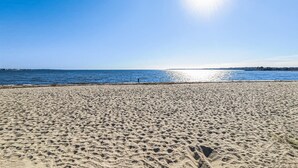 Ein Sandstrand mit Fußspuren, die zum Wasser führen, ein klarer Himmel und eine entfernte Küstenlinie.