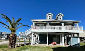 A two-story house with a balcony, a covered porch, and a palm tree in the foreground. - Rockin' Redfish relaxing family beach house, fisherman's paradise. (Crystal Beach)