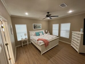 Een slaapkamer met een bed, nachtkastje, dressoir, plafondventilator en drie ramen met jaloezieën.