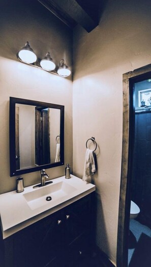 Un baño con lavamanos blanco, muebles oscuros, un espejo con marco de madera y una toalla colgada en un aro.