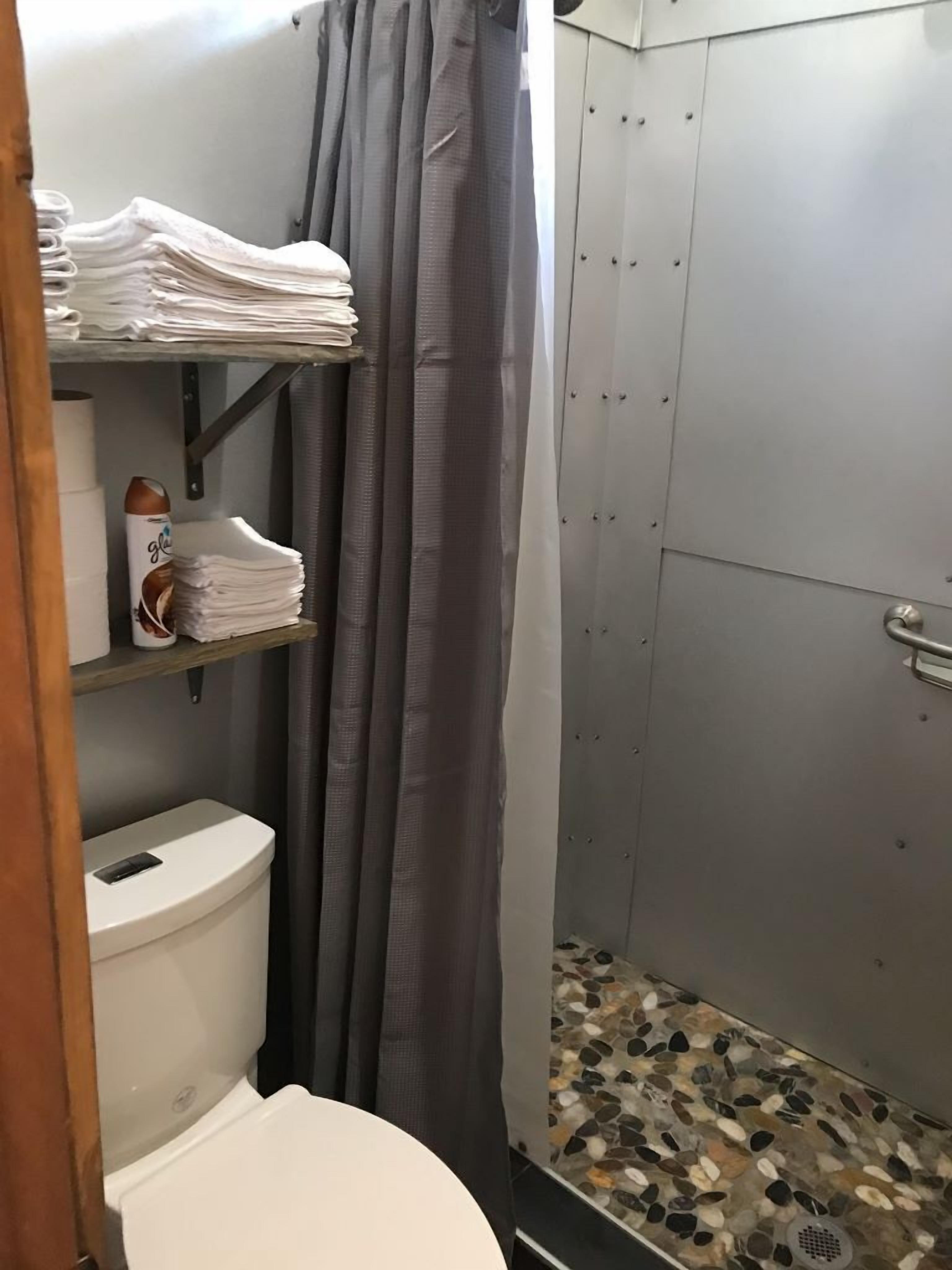 Ein kleines Badezimmer mit Toilette, einer Dusche mit Kieselboden sowie Regalen mit gefalteten Handtüchern und einer Flasche.
