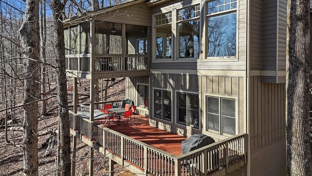 A two-story wooden house with a deck, large windows, and a view of the surrounding trees.