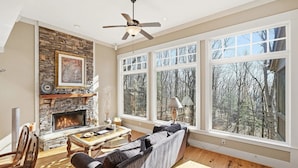 A cozy living room with a fireplace, large windows, and a ceiling fan.