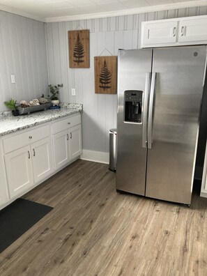 A modern kitchen with white cabinets, a stainless steel refrigerator, a water dispenser, and wooden wall decor. - Farmhouse with game room, creek and fire pit (Johnson City)