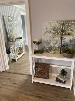 A hallway with a white console table displaying a painting, a vase with flowers, and a basket of green apples. - Farmhouse with game room, creek and fire pit (Johnson City)