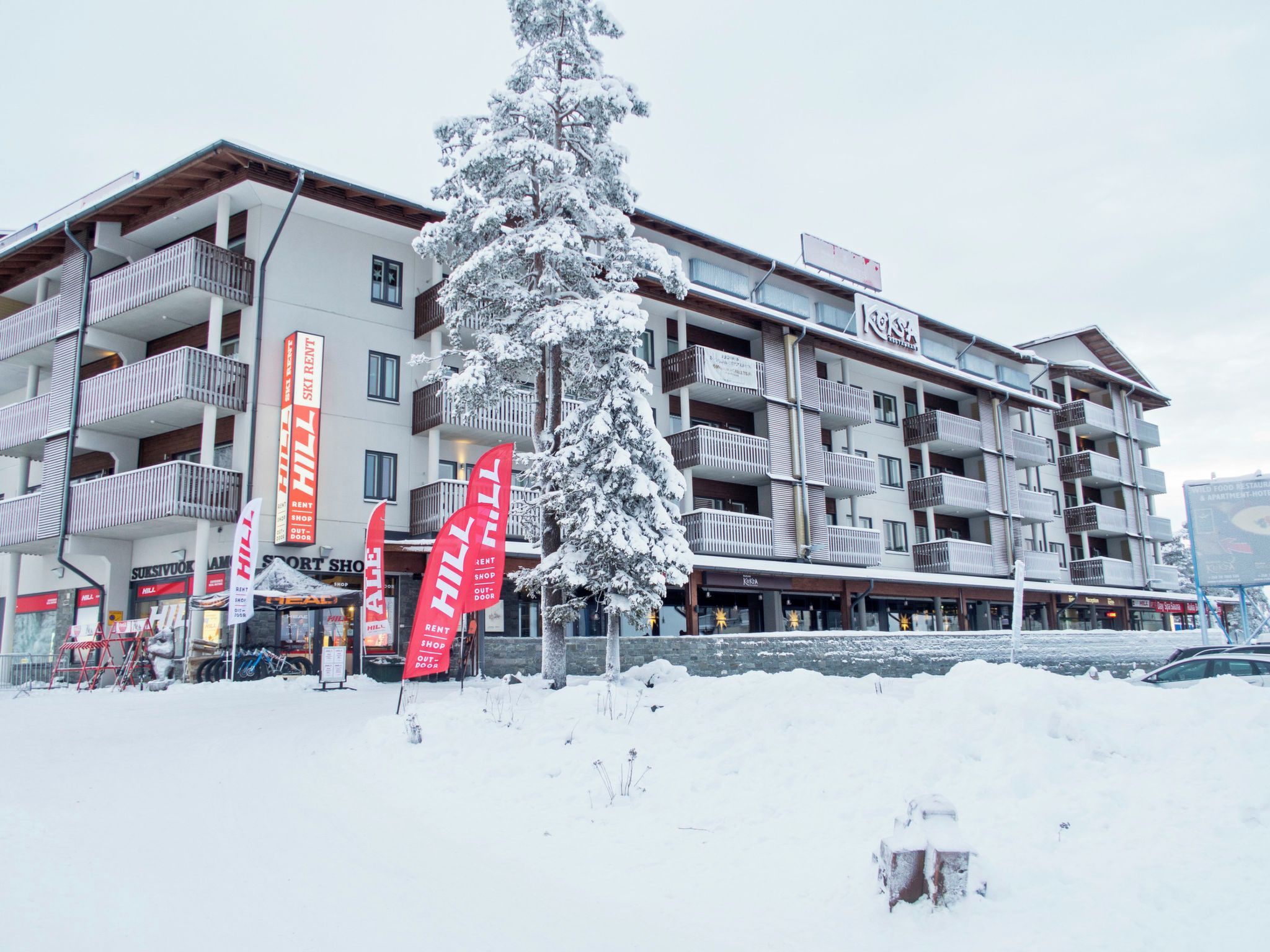 Un hôtel à plusieurs étages, doté de balcons, de panneaux indiquant un service de location de skis et d’un arbre enneigé devant l’entrée.