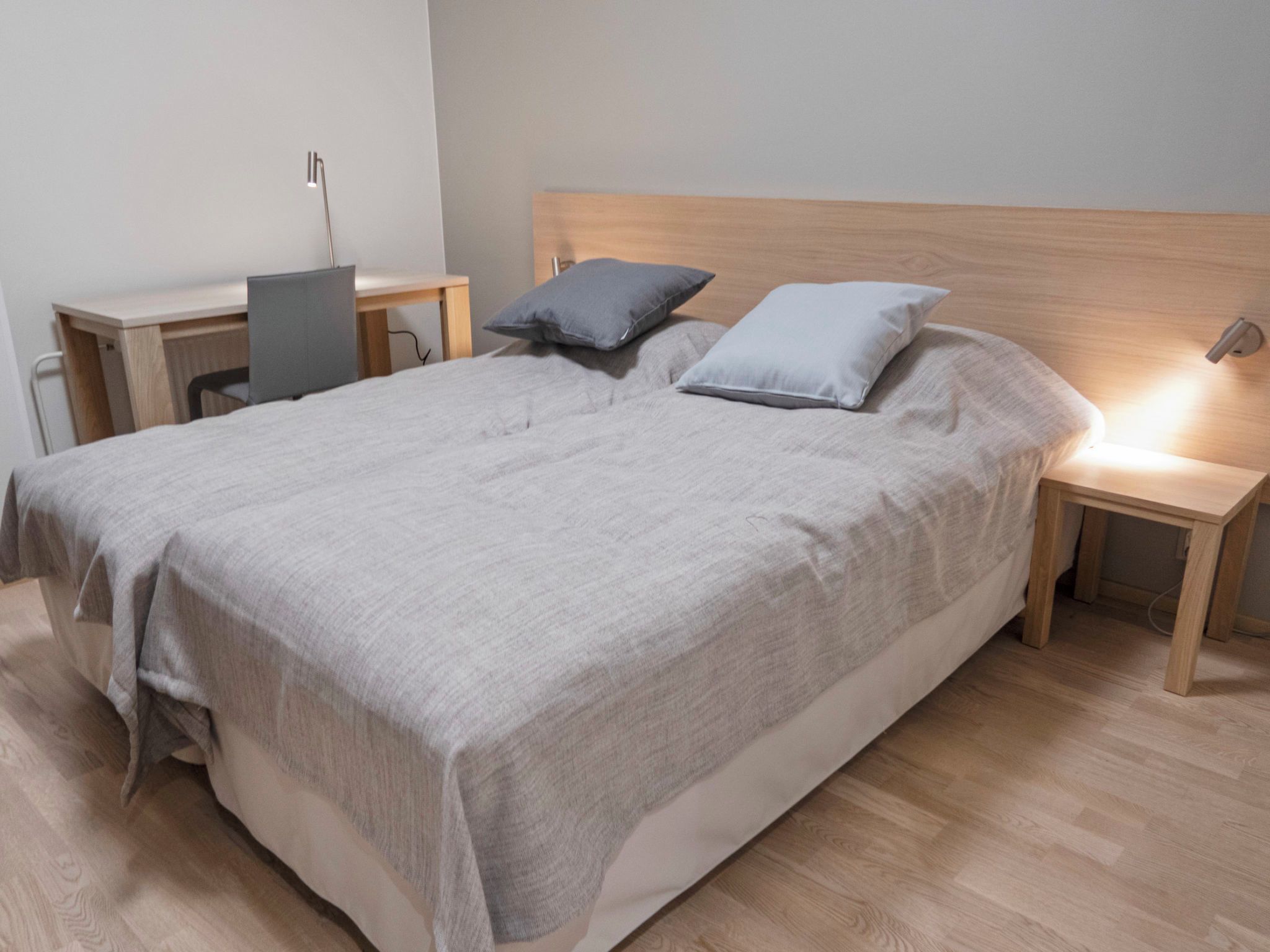 A neatly made bed with a grey blanket, two pillows, and a wooden headboard. There are two bedside tables with lamps, one on each side of the bed.