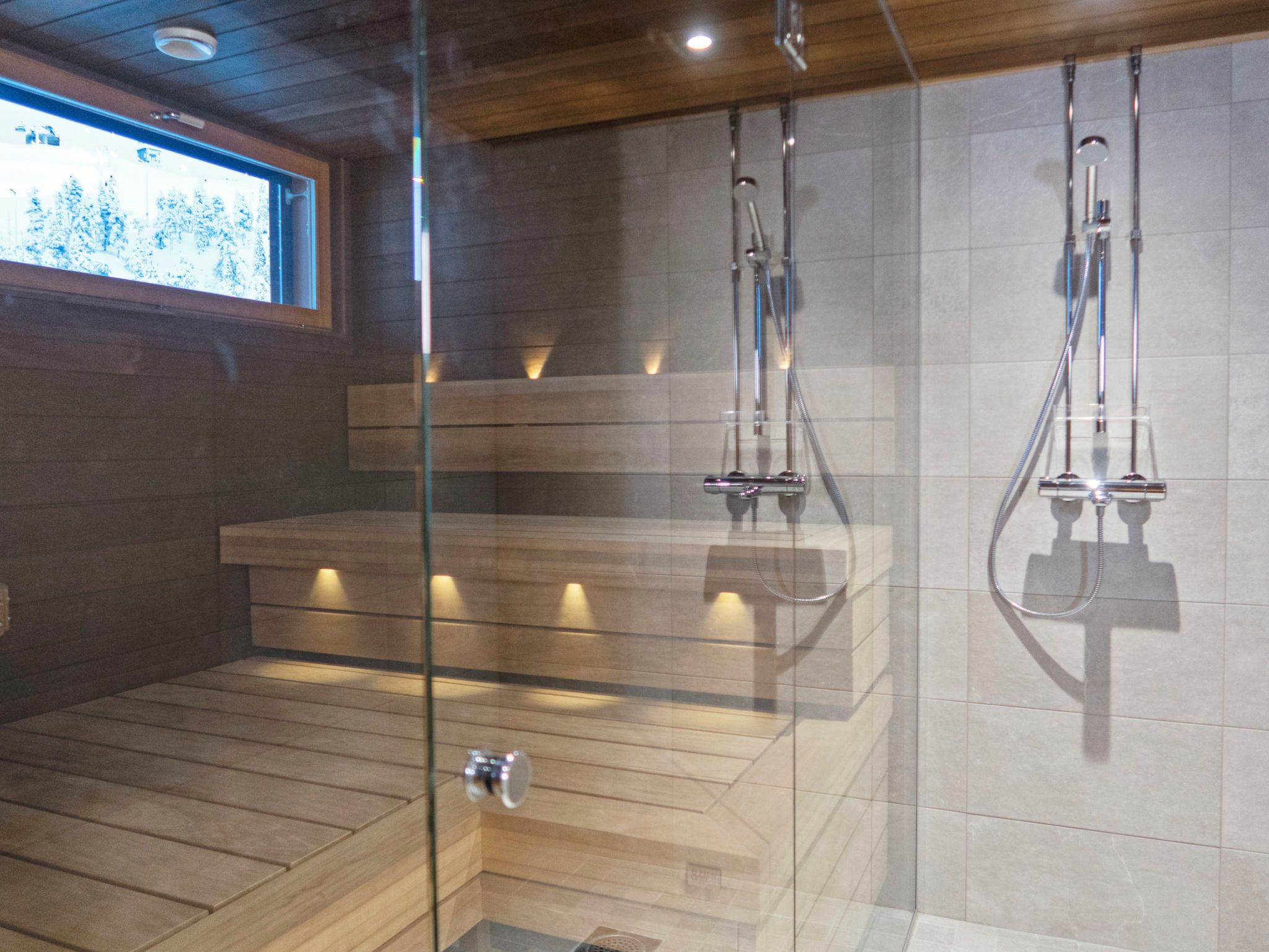 A modern shower area with glass doors, wooden benches, and a tiled wall.